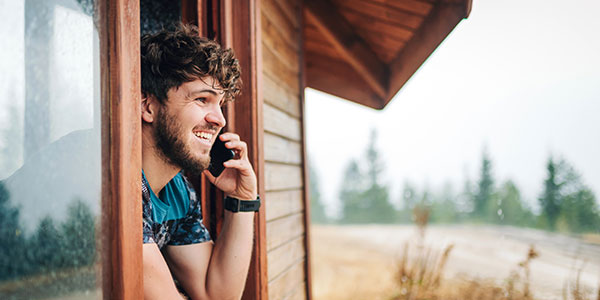A photograph of a young man leaning out a cottage window with a cell phone to his ear.